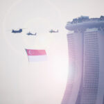Fine art image of a Chinook helicopter flying across Marina Bay Sands with the Singapore flag, escorted by two Apache helicopters in the evening sky.