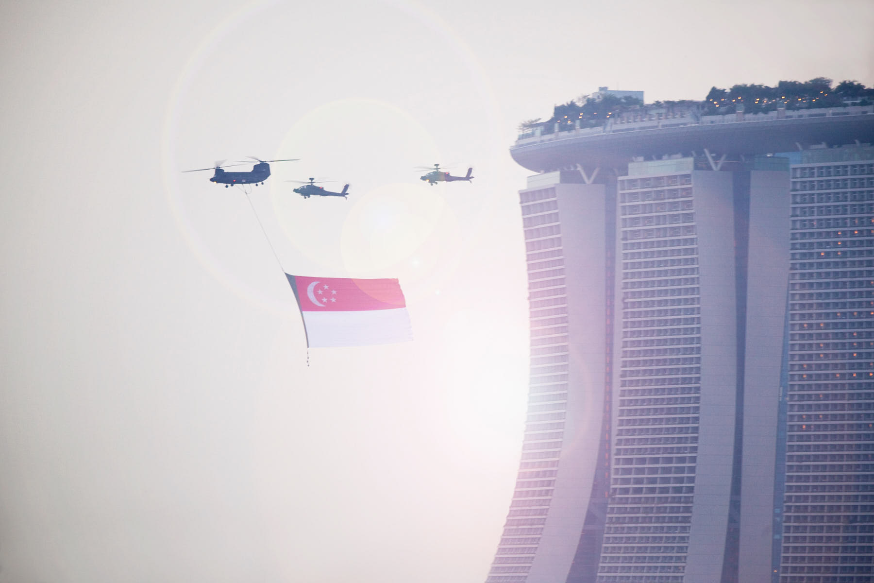 Fine art image of a Chinook helicopter flying across Marina Bay Sands with the Singapore flag, escorted by two Apache helicopters in the evening sky.