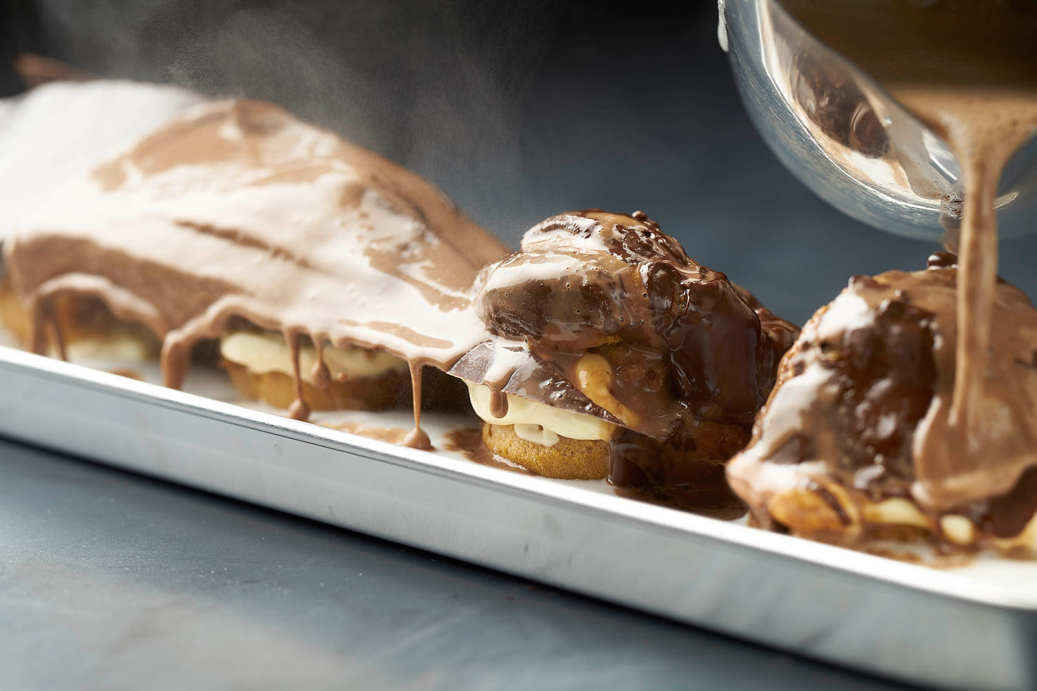 Close-up action shot of a dessert with rich chocolate sauce pouring over it, accompanied by rising steam.